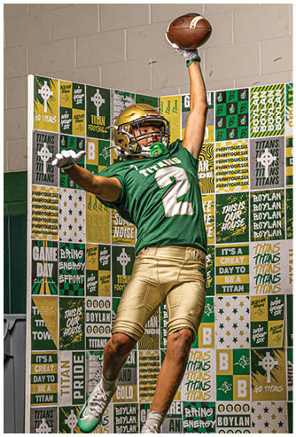 A person wearing a green and gold football uniform is jumping in the air with one arm extended upward holding a football against a backdrop covered in green, gold, and white sports-themed signs and slogans. Description generated by AI