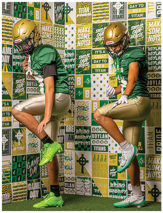 Two people wearing green football jerseys, gold helmets, and gold pants are posing with one leg raised against a colorful wall covered in sports-themed text and graphics. Description generated by AI