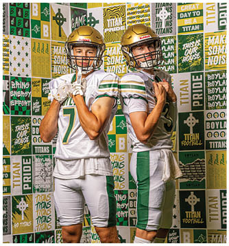 Two adults wearing gold football helmets and white jerseys with green and gold accents pose back to back in front of a colorful wall covered with sports-themed posters and slogans. Description generated by AI