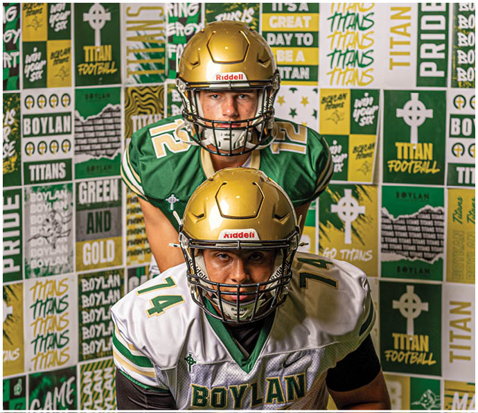 Two people wearing football helmets and Boylan football jerseys pose in front of a wall covered with Boylan Titans-themed posters and banners. Description generated by AI