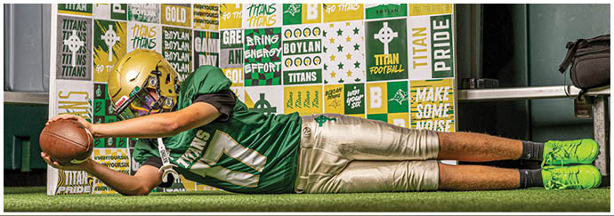 A person wearing a green football jersey and gold pants is lying on the ground reaching out to catch a football in front of a colorful wall covered with sports-themed posters. Description generated by AI