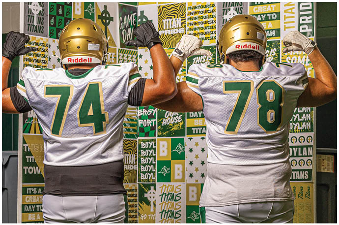 Two people wearing football uniforms and helmets stand with their backs to the camera, flexing their arms in front of a colorful wall covered with sports-themed posters and slogans. Description generated by AI