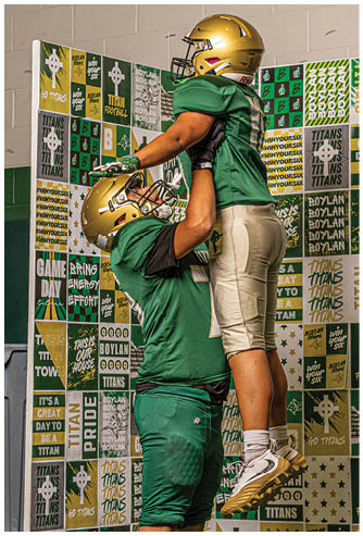 Two people wearing green and gold football uniforms are celebrating with one person lifting the other in front of a backdrop covered in sports-themed signs and slogans. Description generated by AI