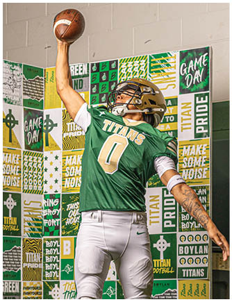 A person wearing a green and gold football uniform with the number zero is reaching up with one hand to catch a football against a backdrop covered in green, gold, and white sports-themed posters. Description generated by AI