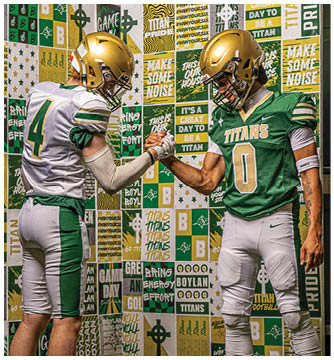 Two people wearing football helmets and jerseys are clasping hands in front of a colorful wall covered with motivational sports-themed posters. Description generated by AI