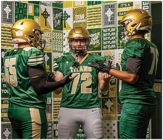 Three people wearing green and gold football uniforms and helmets stand facing each other in front of a wall covered with green, gold, and white sports-themed posters. Description generated by AI