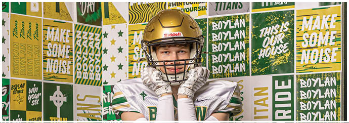 A person wearing a white football jersey with green and gold accents stands with crossed arms above a gold football helmet in front of a green and gold backdrop featuring the words Boylan, Titan Football, and Titans. Description generated by AI