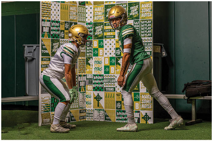 Two people wearing green and white football uniforms and gold helmets are positioned facing each other on an indoor turf field with a colorful backdrop behind them. Description generated by AI