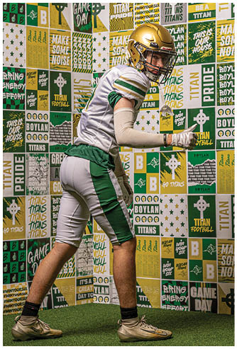 A person wearing a green and white football uniform with a gold helmet is posed in a ready stance against a wall covered in green, gold, and white sports-themed posters and banners. Description generated by AI