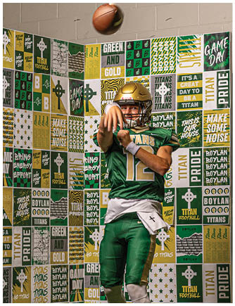 A person wearing a green football uniform with the number 12 is jumping and preparing to throw a football in front of a colorful backdrop covered with motivational sports phrases and team logos. Description generated by AI