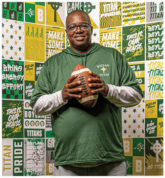 A person wearing glasses and a green Boylan Titans shirt is holding a football and standing in front of a colorful wall covered with Boylan Titans and motivational sports-themed posters. Description generated by AI