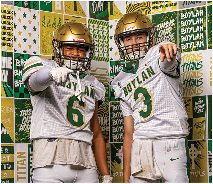 Two people wearing football uniforms and helmets stand in front of a colorful backdrop with sports-themed text and graphics, both pointing toward the camera. Description generated by AI