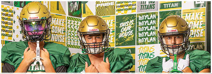Three people wearing green football jerseys and gold helmets stand in front of a wall covered with motivational sports-themed posters. Description generated by AI