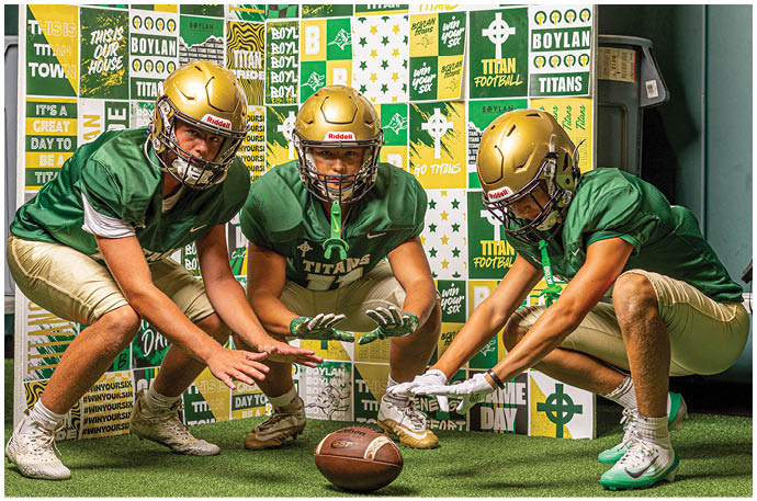 Three people wearing green football jerseys and gold helmets are crouched down in a football stance inside a room decorated with sports-themed posters and banners. Description generated by AI