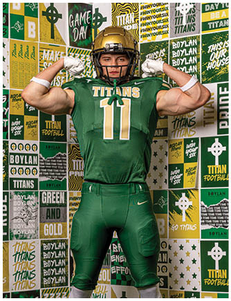 A person wearing a green football jersey and gold helmet flexes their arm muscle in front of a colorful backdrop with football-related text and logos. Description generated by AI