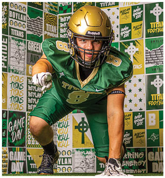 A person wearing a green football uniform and gold helmet is posing in a dynamic stance in front of a wall covered with various sports-themed posters and signs. Description generated by AI