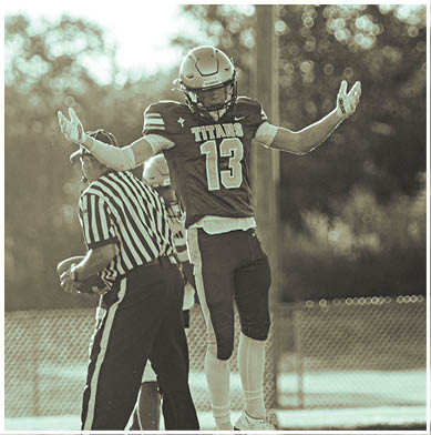 A person wearing a football uniform with the number 13 and the word Titans on the jersey is standing with arms raised next to a referee holding a football on a field. Description generated by AI