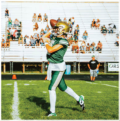 An adult in a green and white football uniform with a gold helmet is catching a football on a grassy field with empty bleachers and scattered spectators in the background. Description generated by AI