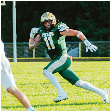 A person in a green football uniform with the number 11 is running toward another person in a white football uniform holding a football on a grassy field. Description generated by AI