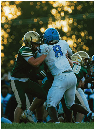Two people wearing football helmets and uniforms are engaged in a close physical play on a field with blurred lights in the background. Description generated by AI
