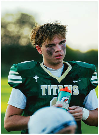 A person with messy hair and black face paint under their eyes is wearing a green sports jersey with white stripes and a cross emblem. Description generated by AI