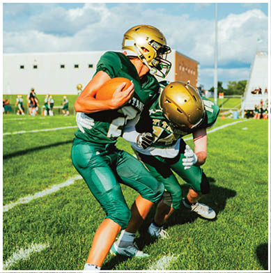 Two people in green football uniforms and gold helmets are engaged in a play on a grassy field with a partly cloudy sky and buildings in the background. Description generated by AI