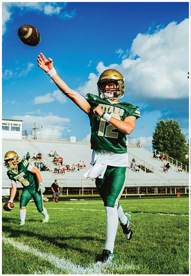 A person wearing a green football uniform and gold helmet is throwing a football on a sunny day with a partly cloudy sky and empty bleachers in the background. Description generated by AI