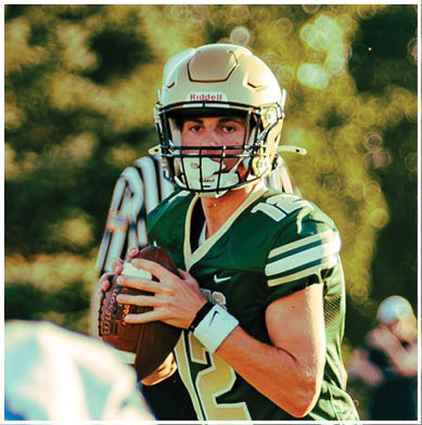 A person wearing a football helmet and green jersey is holding a football and appears to be preparing to throw it on a field with blurred trees in the background. Description generated by AI