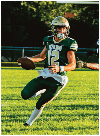 A person wearing a green football uniform with the number 12 and a gold helmet is holding a football and looking to the side on a grassy field with a goalpost and another player reaching toward them. Description generated by AI