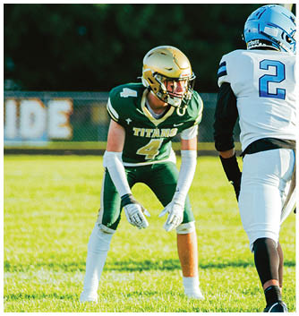 A person wearing a green football uniform with the word Titans and the number 4 is crouched in a ready position facing another person in a white and blue football uniform on a grassy field. Description generated by AI