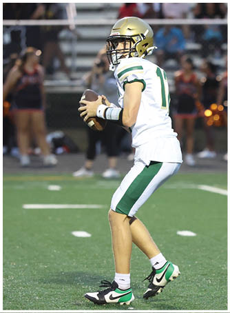 Two people wearing football helmets and uniforms are engaged in a close physical play on a field with blurred lights in the background. Description generated by AI