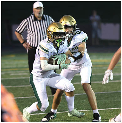 Two people in green football uniforms and gold helmets are engaged in a play on a grassy field with a partly cloudy sky and buildings in the background. Description generated by AI