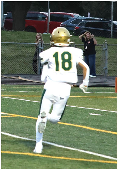 A person wearing a green football uniform and gold helmet is throwing a football on a sunny day with a partly cloudy sky and empty bleachers in the background. Description generated by AI