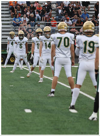 A person wearing a green football uniform with the number 12 and a gold helmet is holding a football and looking to the side on a grassy field with a goalpost and another player reaching toward them. Description generated by AI