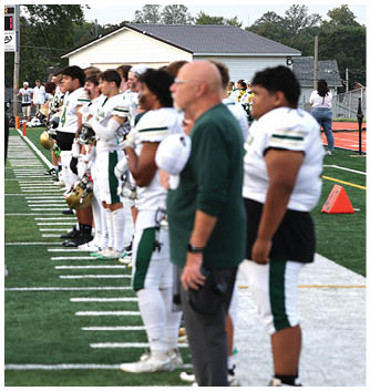 A person wearing a green football uniform with the word Titans and the number 4 is crouched in a ready position facing another person in a white and blue football uniform on a grassy field. Description generated by AI