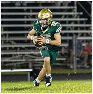 A person in a green football uniform and gold helmet is holding a football and running on a grassy field with another player and a person in a white and red uniform standing in the background near bleachers. Description generated by AI