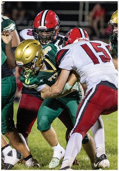 Two people in green and gold football uniforms are tackling a person in a white and red football uniform on a grassy field during a game. Description generated by AI