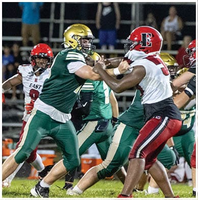 A person in a green and gold football uniform is preparing to throw a football while being closely pursued by two people in red and white football uniforms on a grassy field with blurred spectators in the background. Description generated by AI