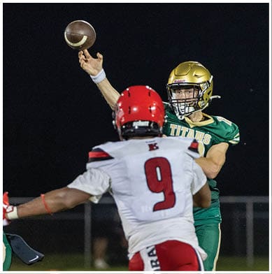 A person in a green football uniform with a gold helmet is throwing a football while another person in a white uniform with a red helmet and the number nine on the back is facing them. Description generated by AI