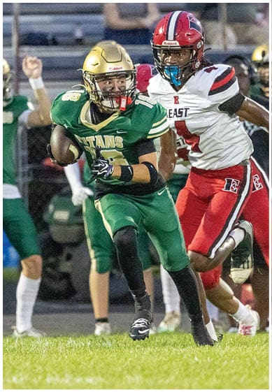 A person in a green football uniform with the word “TITANS" is running with a football while being pursued by a person in a white and red uniform on a grassy field. Description generated by AI