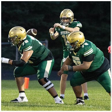 Two people in football uniforms are engaged in a play on a grassy field with one person in green and gold gear preparing to catch or block a football. Description generated by AI