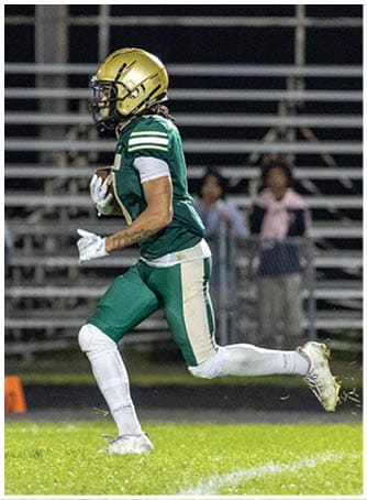 A person wearing a green and gold football uniform is running on a grassy field with blurred spectators in the background. Description generated by AI