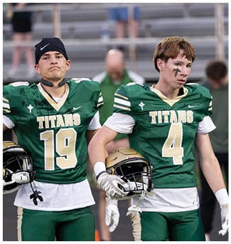Two adults wearing green football uniforms with the word Titans stand side by side on a field holding helmets, with one person having face paint. Description generated by AI