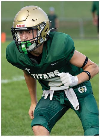 A person wearing a gold football helmet and green jersey is crouched on a grassy field with blurred spectators in the background. Description generated by AI