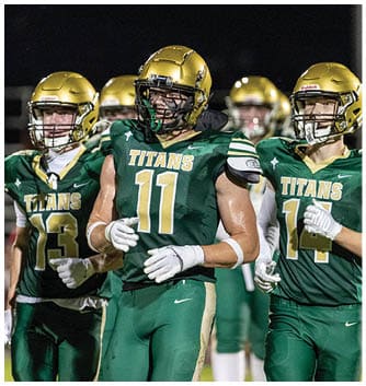 A person wearing a green football uniform with the word “TITANS" and the number 13 on it is standing on a field with other players in the background. Description generated by AI