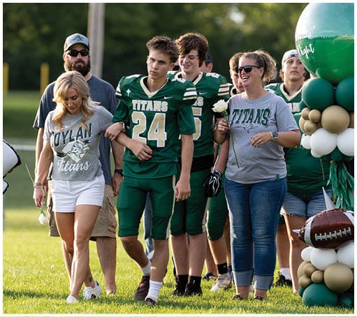 People in green football uniforms and a person in a Titans shirt walk together outdoors near a balloon arrangement and a football. Description generated by AI