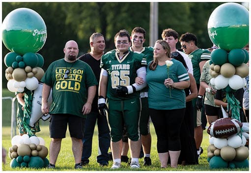 A group of people stands together on a sports field with one person in a football uniform holding a white rose and others around them, next to a balloon arrangement featuring a football. Description generated by AI
