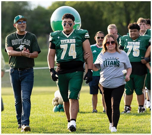 Two people are walking on grass, one wearing jeans and casual shoes and the other wearing a green sports uniform with gloves and cleats. Description generated by AI