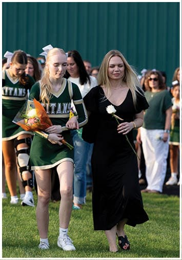 An adult in a black dress holding a white rose walks arm-in-arm with a person in a green cheerleading uniform holding flowers, with several people walking behind them. Description generated by AI