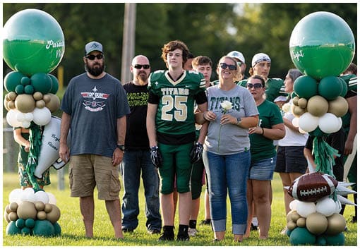 A group of people stand outdoors with one person wearing a green football jersey numbered 25 and others wearing casual clothing including sunglasses and hats. Description generated by AI